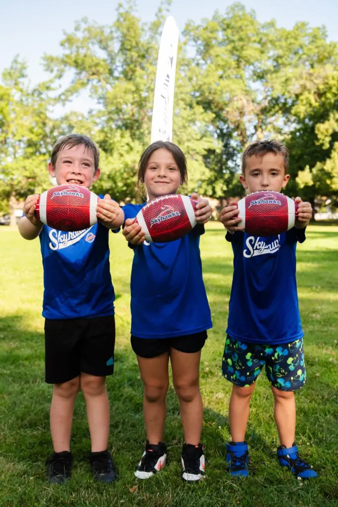 3 young kids in blue jerseys holding small footballs